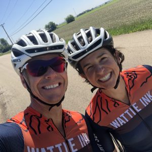 Dr. Bruce Noxon selfie, outdoors in an orange and blue road biking team jersey and helmet with a female teammate who is dressed similarly.