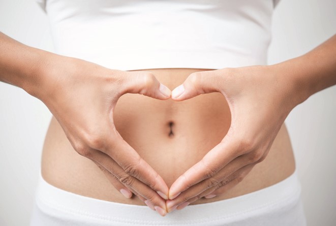 A woman's mid section is pictured. In a white crop top and leggings. Her hands forma heart shape around her belly button.